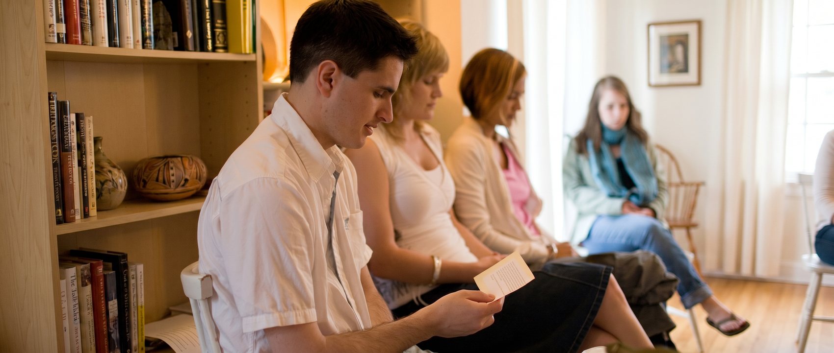 A young man reads a prayer aloud as friends listen during a devotional gathering in a sunlit, book-lined room